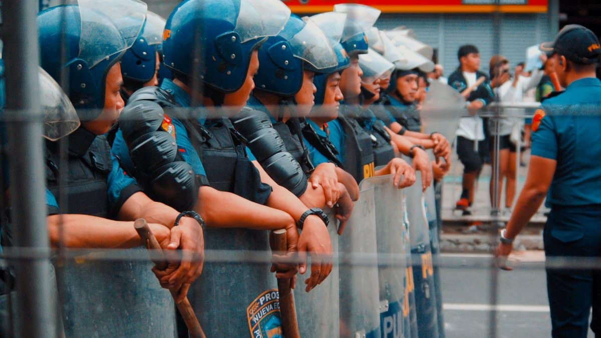 A line of police officers in riot gear standing on a street during a public gathering.