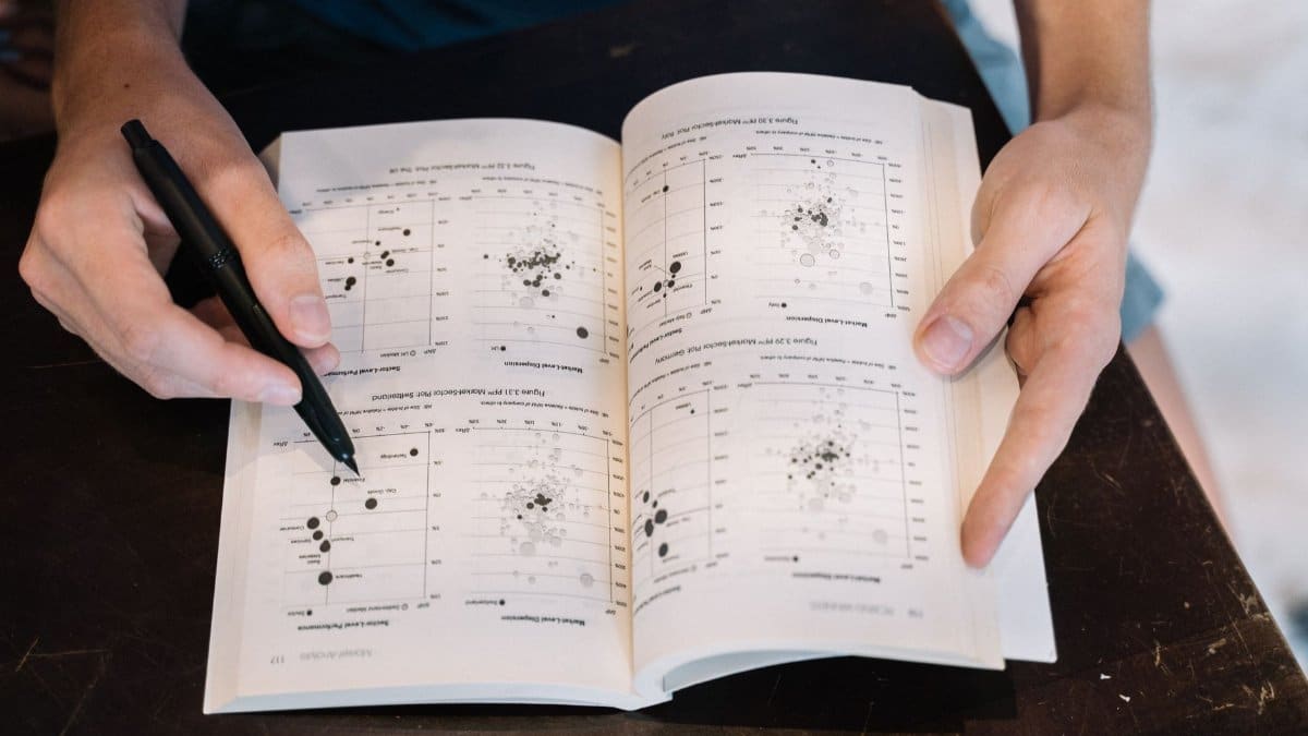 Person analyzing data charts in a book using a pen, close-up perspective.