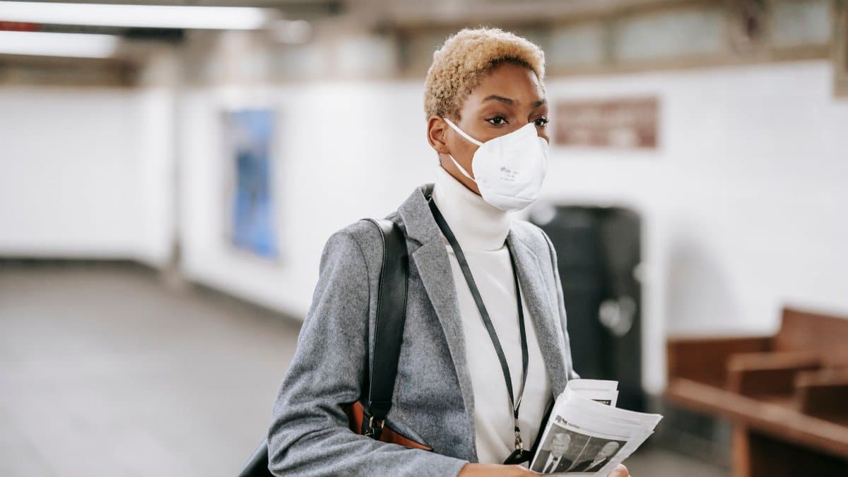 Thoughtful young African American female in formal wear and face mask standing with newspaper in hands in public hallway and looking away