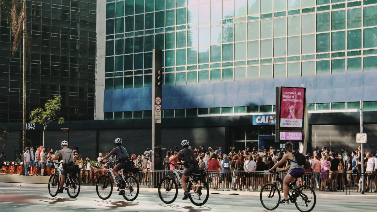 Cyclists ride past a crowded building entrance in a sunny urban setting, showcasing city life and mobility.