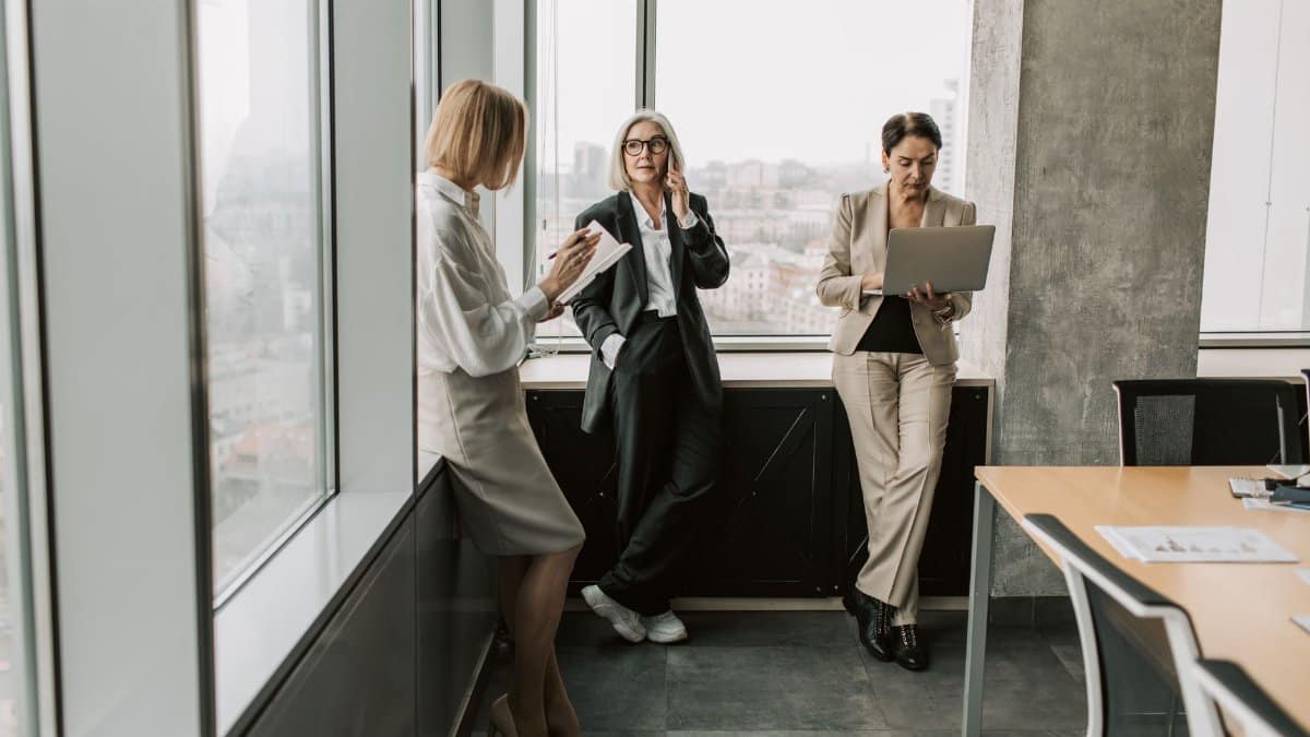 Three women in office attire collaborating near large windows, using technology.