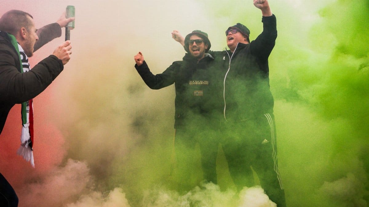 Group of sports fans celebrating with colorful smoke outdoors, showing excitement and joy.