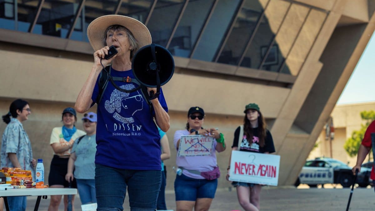 A group at a protest with an elderly woman leading through a megaphone outdoors.