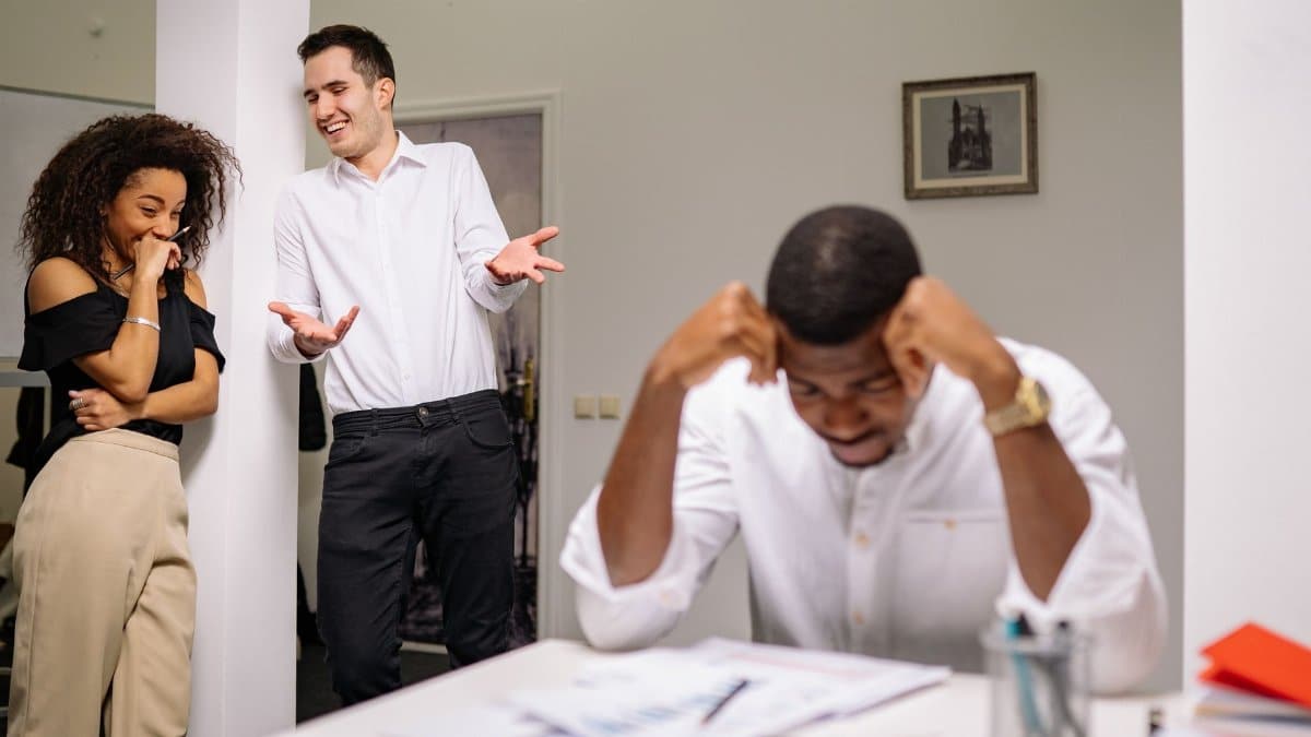 Colleagues sharing a light moment while another looks stressed at his desk in a modern office.