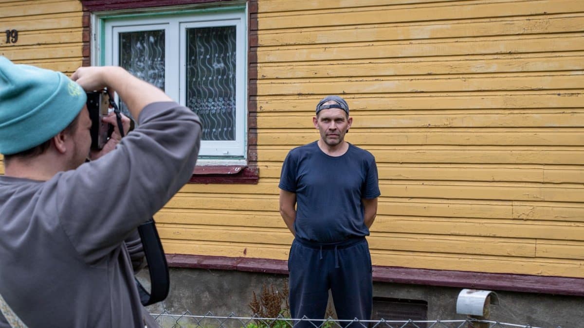 Man posing in front of a rural Estonian wooden house, captured outdoors.