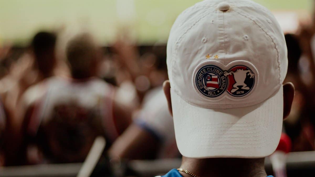 A dedicated football fan sport ing an Esporte Clube Bahia cap watches the game.