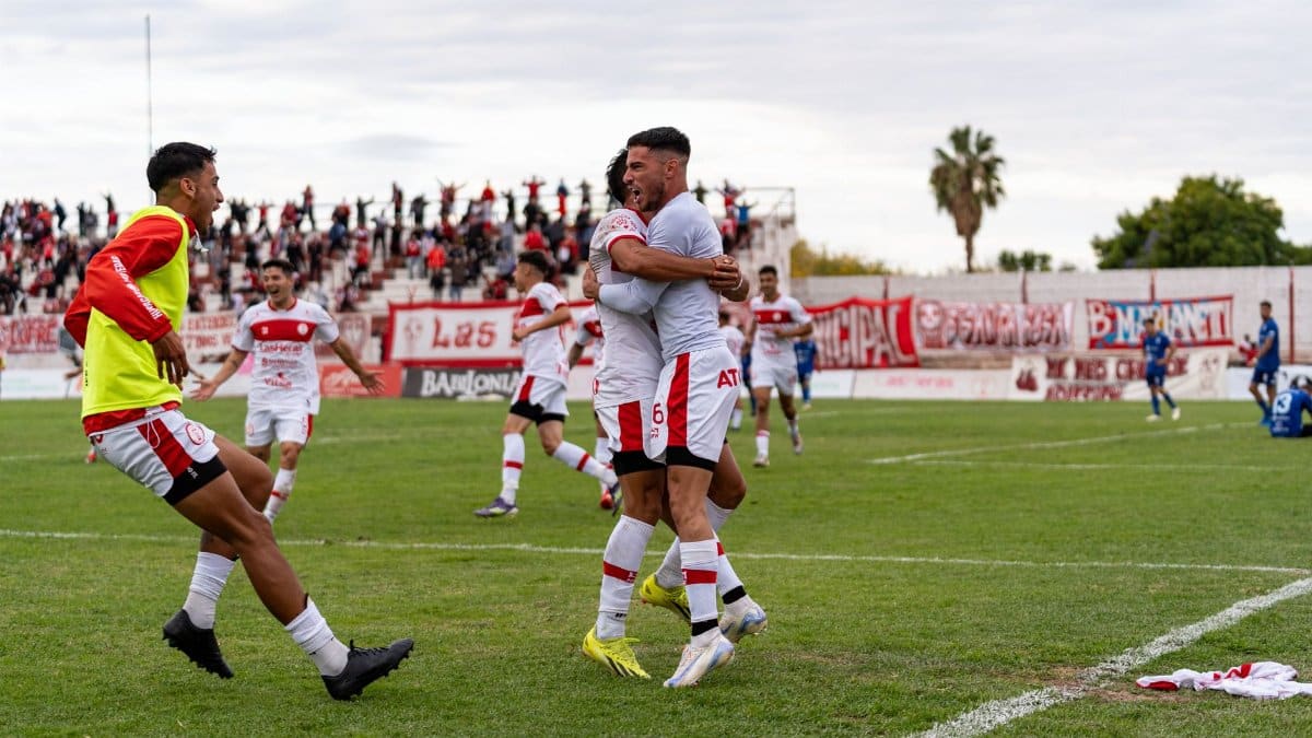Football players celebrating a goal on the field with enthusiastic fans in the background.