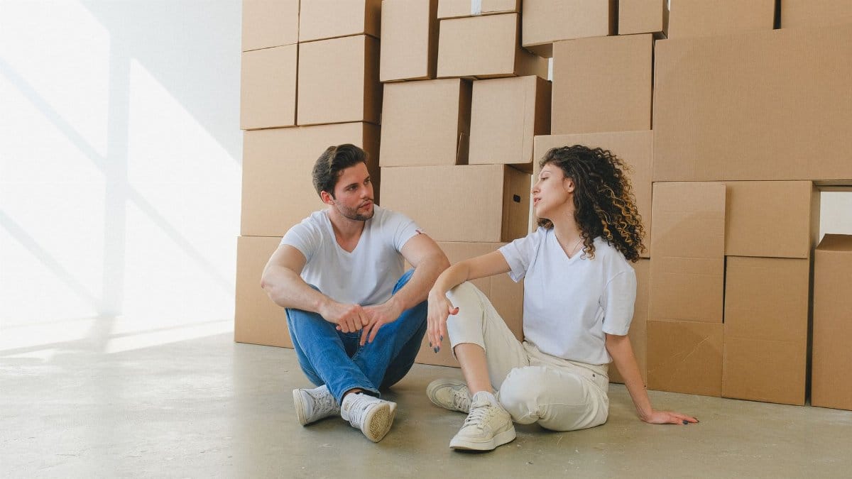 Woman talking to boyfriend with crossed legs sitting on floor against piles of carton containers after moving into house