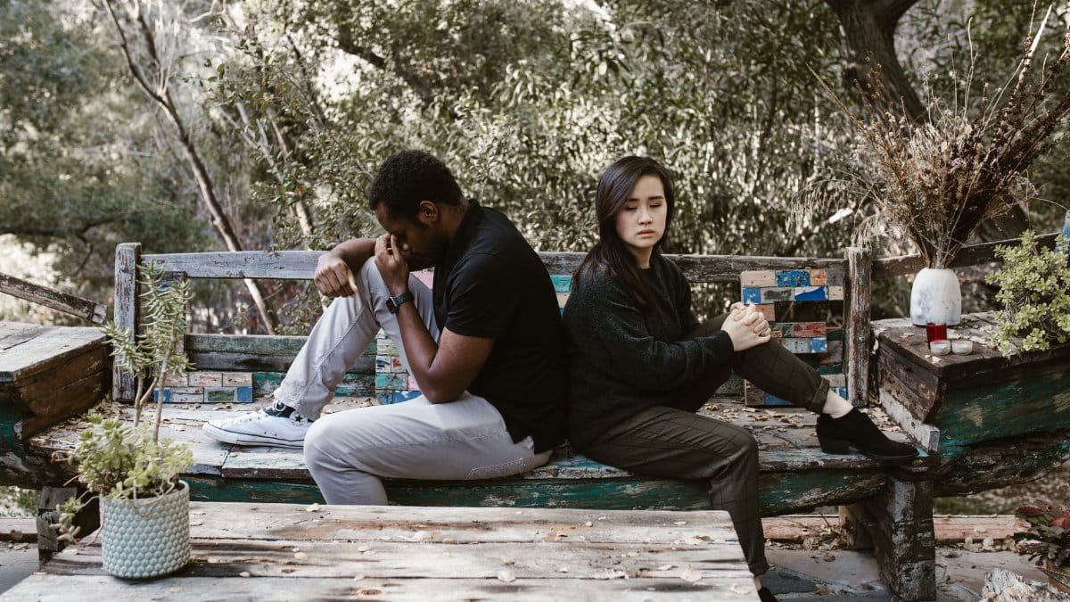 A couple sits back to back on a wooden bench, indicating a disagreement or breakup.