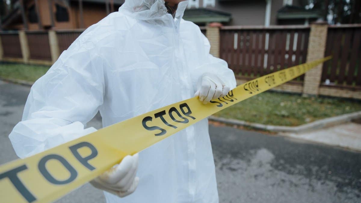 A person wearing a protective suit, handling yellow tape with 'STOP' in an outdoor urban setting.