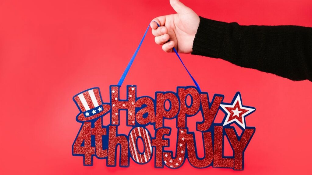A hand holding a glittery 'Happy 4th of July' sign against a vibrant red background.