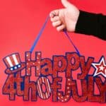 A hand holding a glittery 'Happy 4th of July' sign against a vibrant red background.