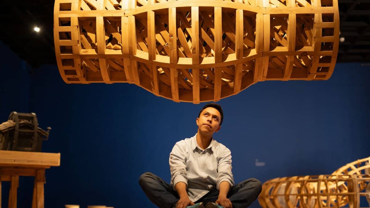 A thoughtful man sits beneath a large abstract wooden sculpture in an art gallery.