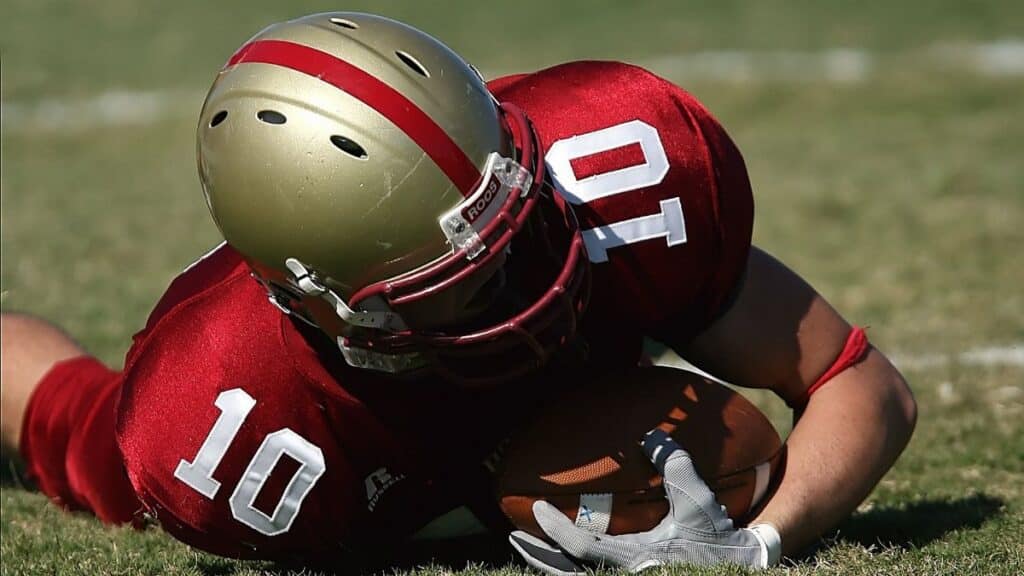 American football player wearing red uniform holding ball on grassy field during day game.