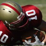 American football player wearing red uniform holding ball on grassy field during day game.