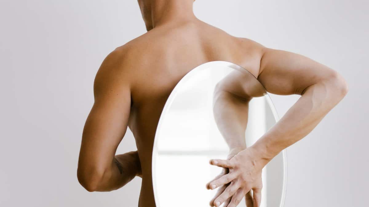 A man in a minimalist setting holding a round mirror reflecting his back.