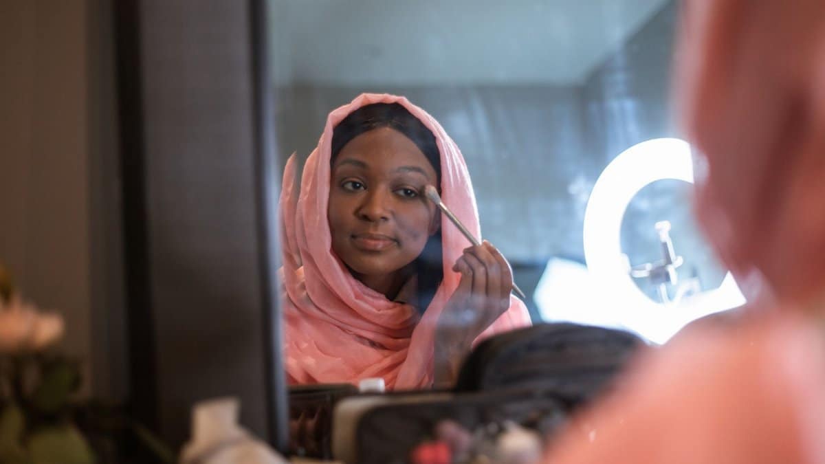 A woman in a hijab applies makeup, reflecting elegance and style in front of a mirror.