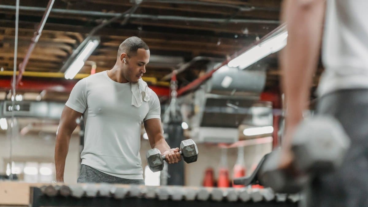 Crop strong young male holding heavy dumbbells while pumping biceps in front of mirror in contemporary fitness center
