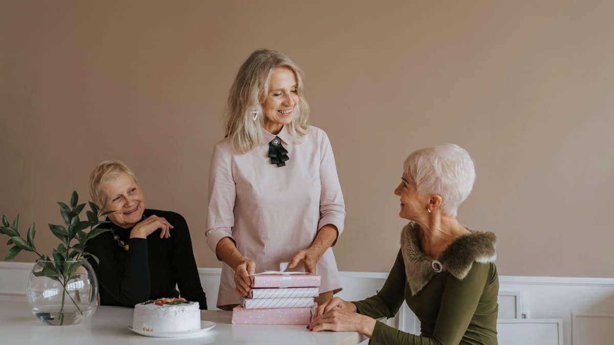 Three senior women celebrate a birthday indoors with gifts and cake, sharing laughter and happiness.