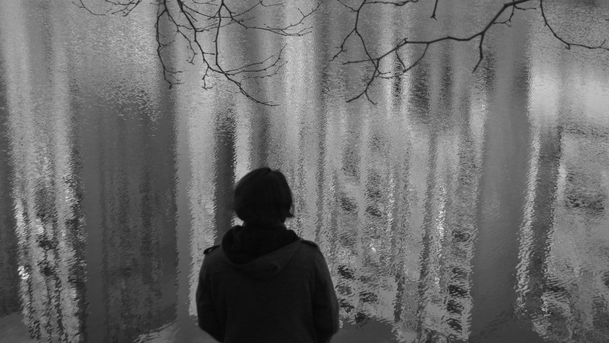 A solitary figure observes urban reflections on a tranquil water surface.