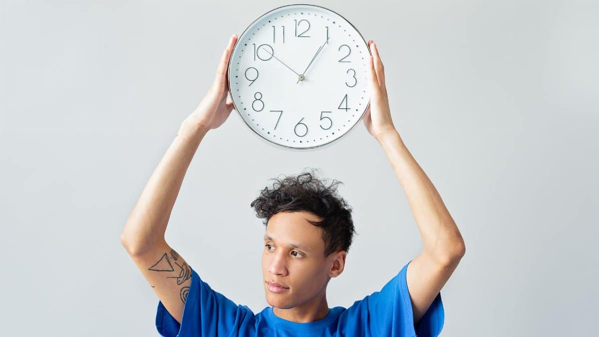 A man in a blue shirt holding a wall clock above his head, contemplating time.