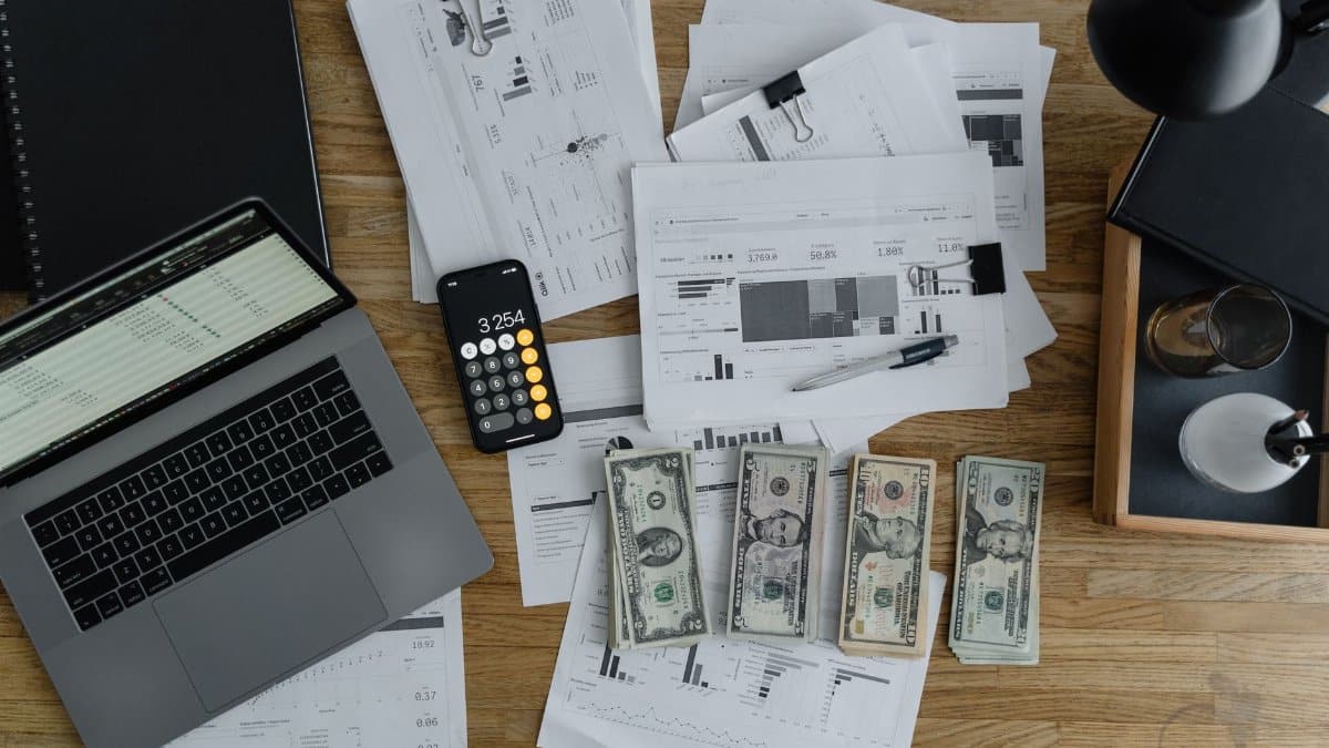 Overhead view of financial documents, cash, and technology on a wooden desk.