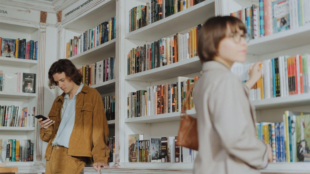 Two people browsing books in a cozy bookstore. A man checks his phone while a woman selects a book.