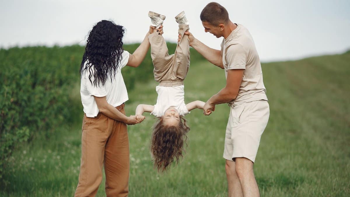 A joyful family enjoys outdoor play, lifting their child upside down in a green field.