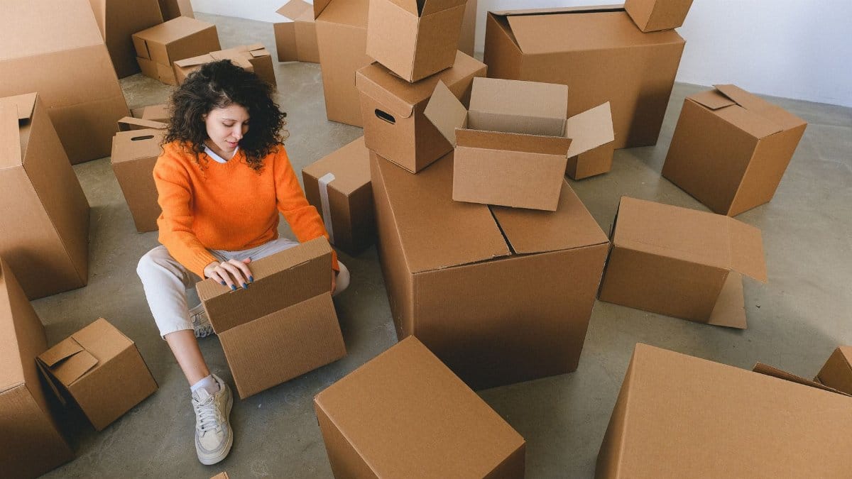 From above of concentrated young female with curly hair in stylish clothes sitting on floor and packing belongings into cardboard boxes before relocation into new apartment