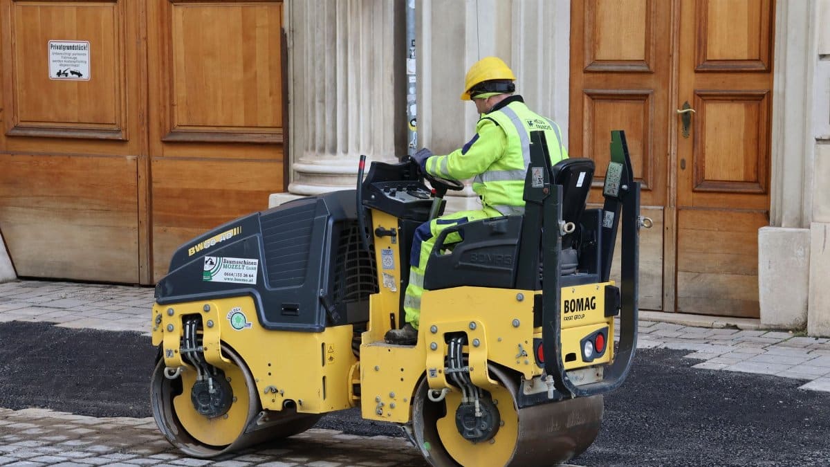 A construction worker in safety gear operates a road roller on a cobblestone street near a historical building.