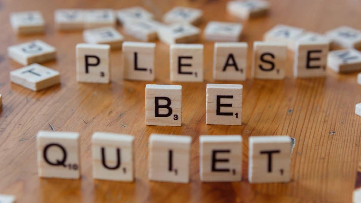 Scrabble tiles on a wooden table spell out 'Please Be Quiet'.