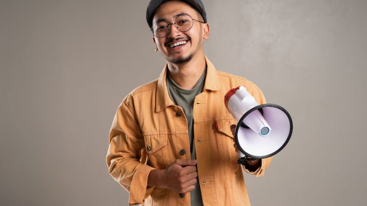 Asian man with eyeglasses and cap smiling while holding a megaphone in a studio shoot.