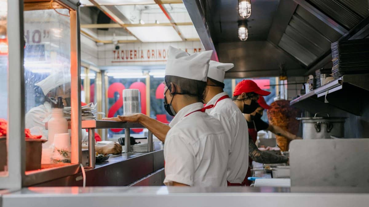 Culinary staff wearing uniforms and masks preparing meals in a bustling restaurant kitchen setting.