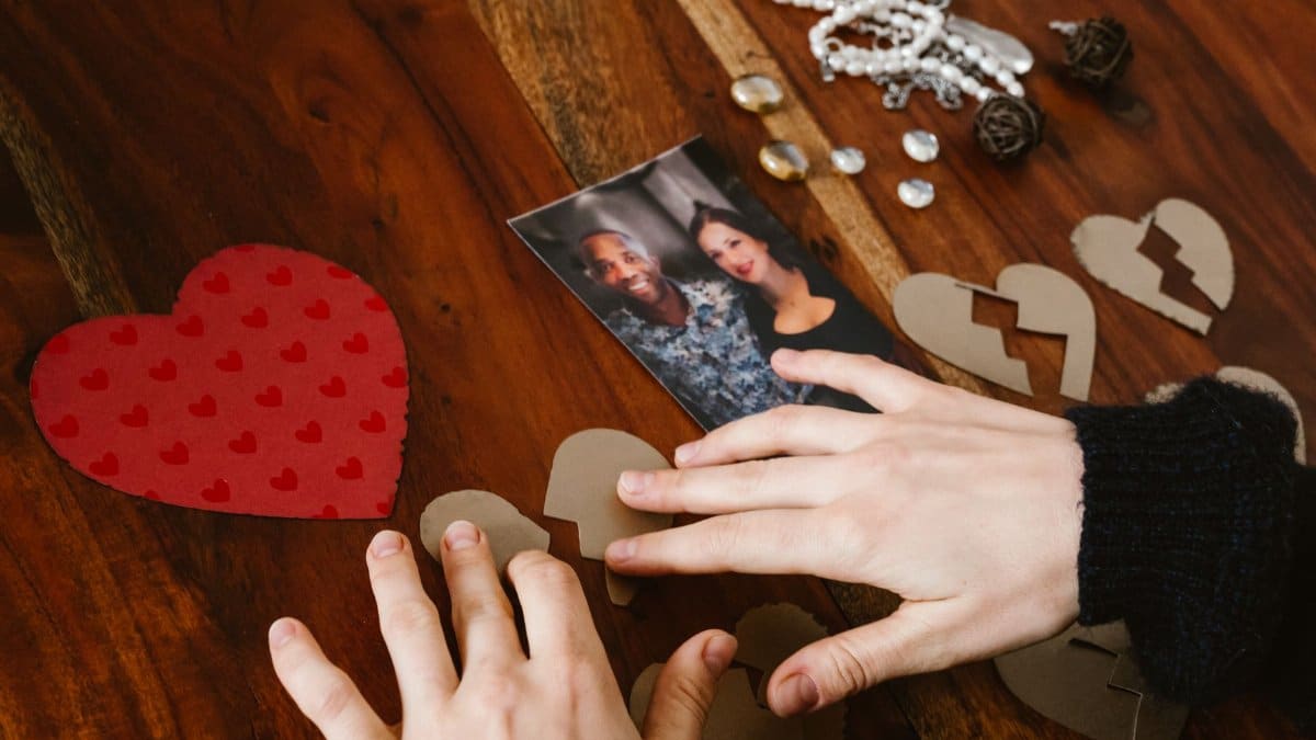 Close-up of hands arranging broken hearts near a couple's photo on a wooden table.