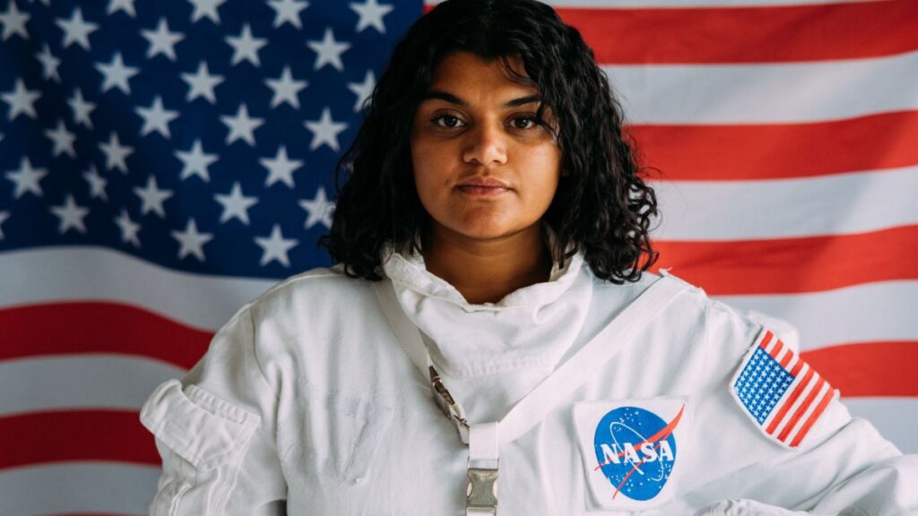 A woman in a NASA astronaut suit poses confidently against an American flag background.