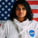 A woman in a NASA astronaut suit poses confidently against an American flag background.