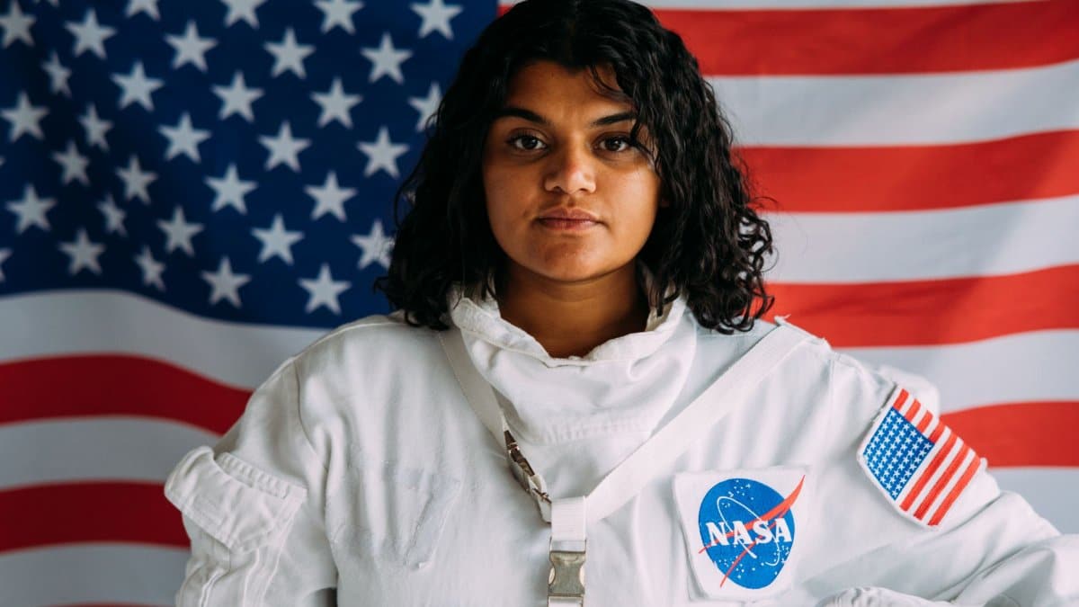 A woman in a NASA astronaut suit poses confidently against an American flag background.