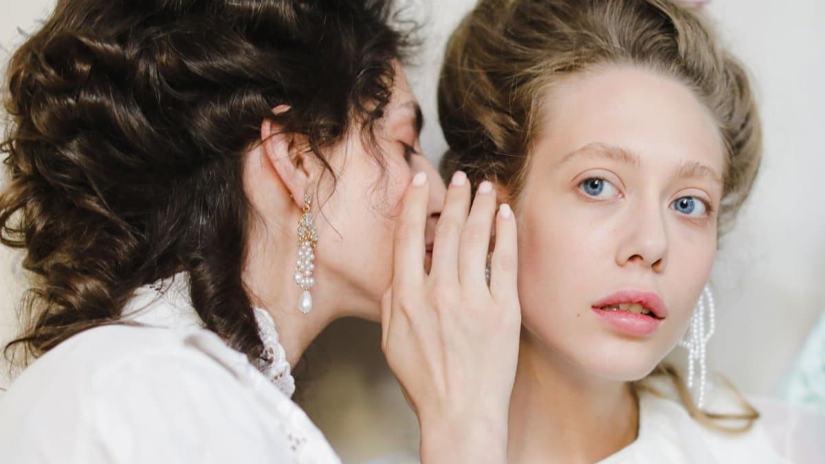 Two women dressed in 19th century fashion share a secret, showcasing elegance and friendship.