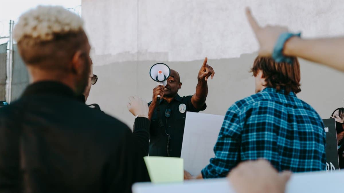 A protest scene with a police officer uses a megaphone to address the gathered crowd during daytime outdoors.