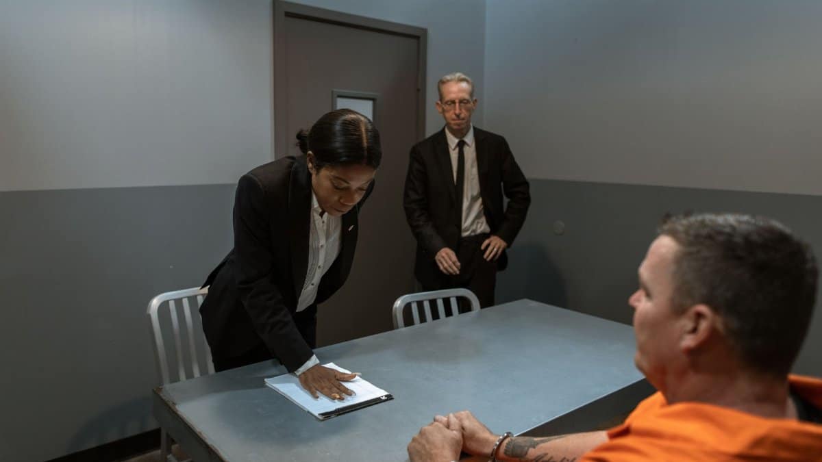 A prisoner in handcuffs is questioned by a detective and lawyer in an interrogation room.
