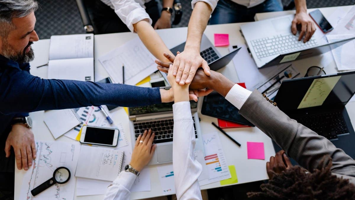 High-angle shot of diverse hands together over a desk with laptops and paperwork, symbolizing teamwork.