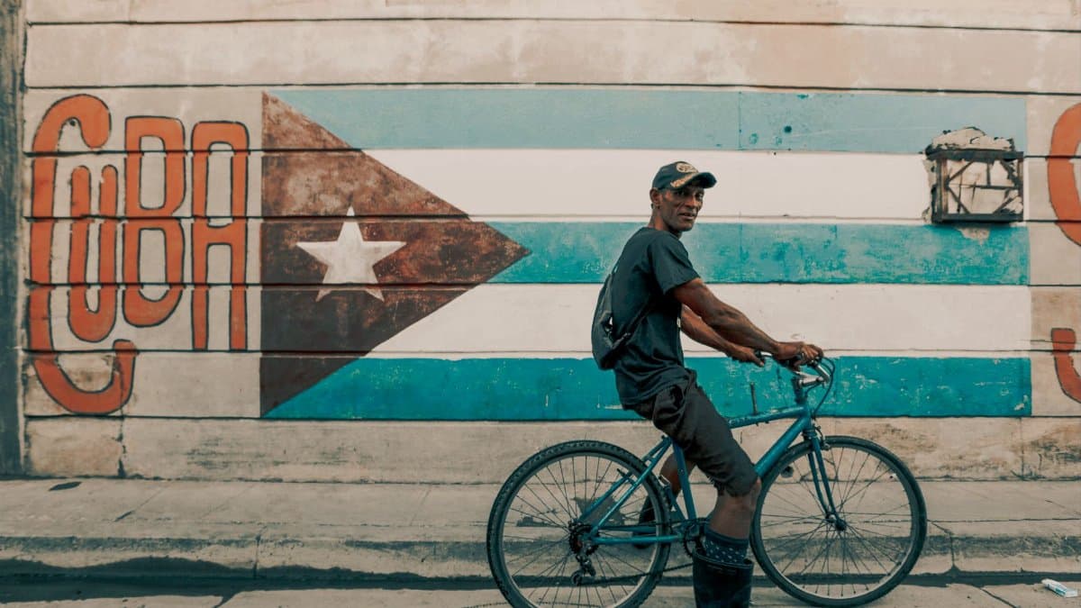 A man rides a bicycle by a vibrant Cuban flag mural in Havana.