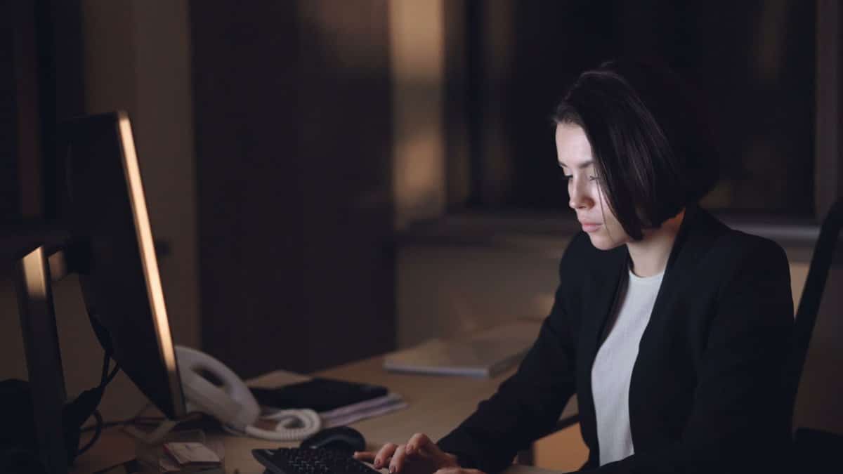 A woman in a suit works late at a dimly lit office desk, focused on her computer.