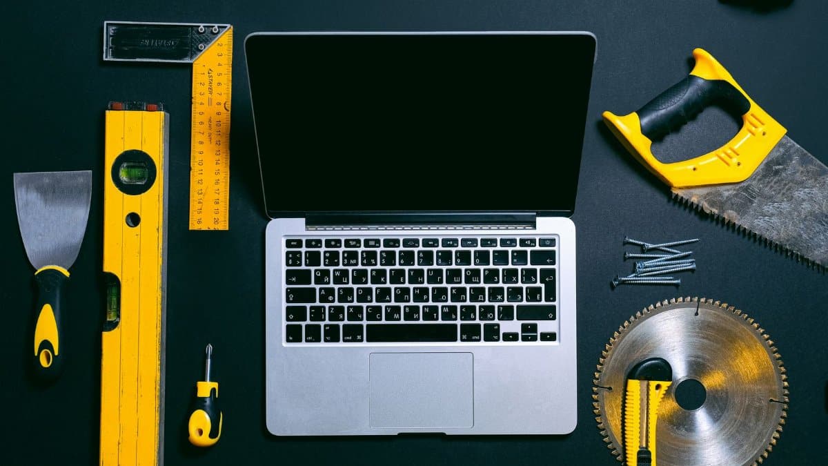 Overhead view of a laptop surrounded by various hand tools on a dark background, representing technology and construction.