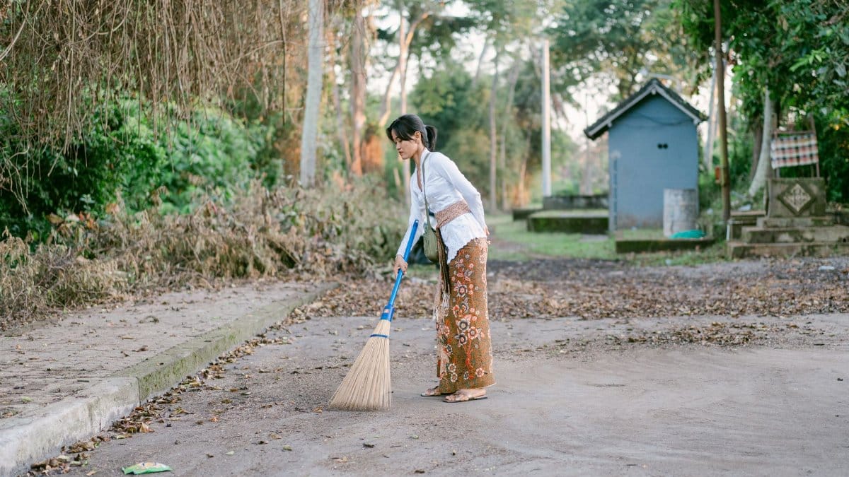 Woman cleaning a street with a broom, surrounded by trees and a serene environment.