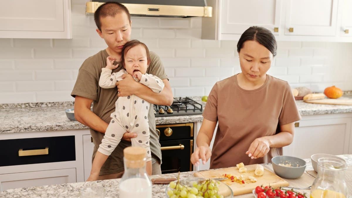 Asian family preparing breakfast in a cozy kitchen setting, with dad holding a baby.