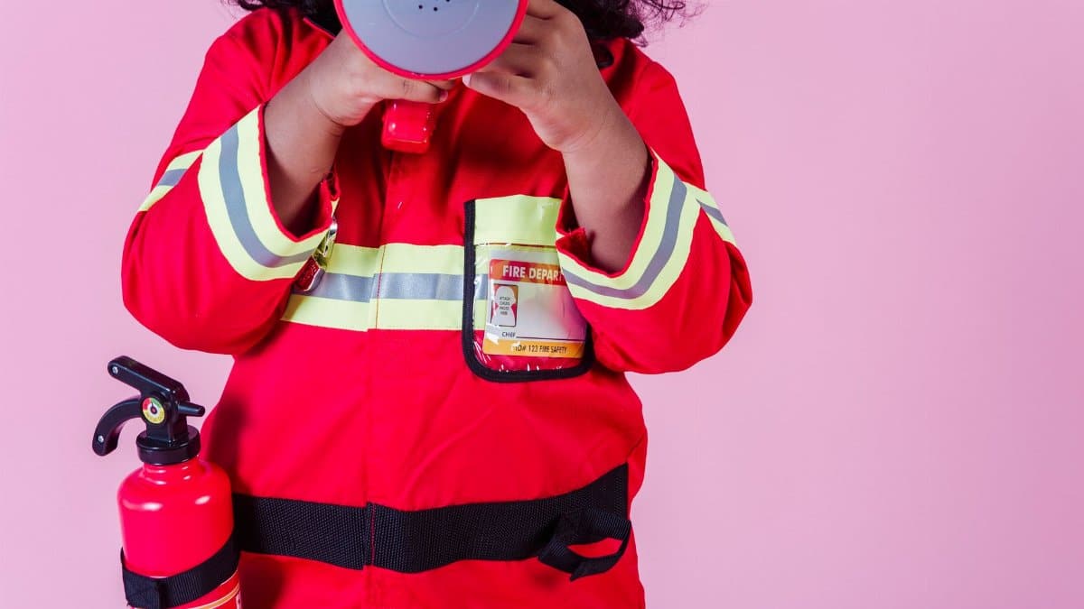 Crop unrecognizable little ethnic kid wearing fireman costume and fire extinguisher while using loudspeaker toy and standing on pink background
