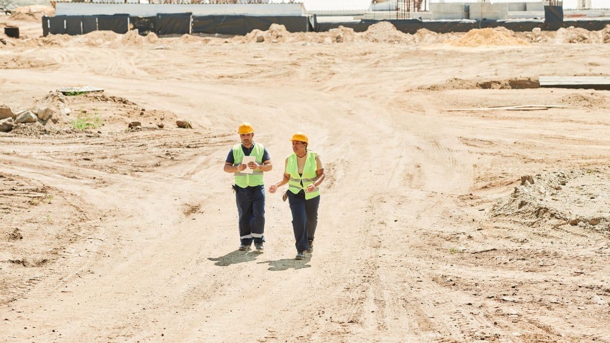 Two workers in safety gear walking across a construction site under a clear sky.