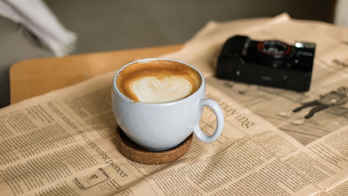 A steamy cup of latte with art sits atop a newspaper beside a vintage camera, evoking a cozy ambiance.