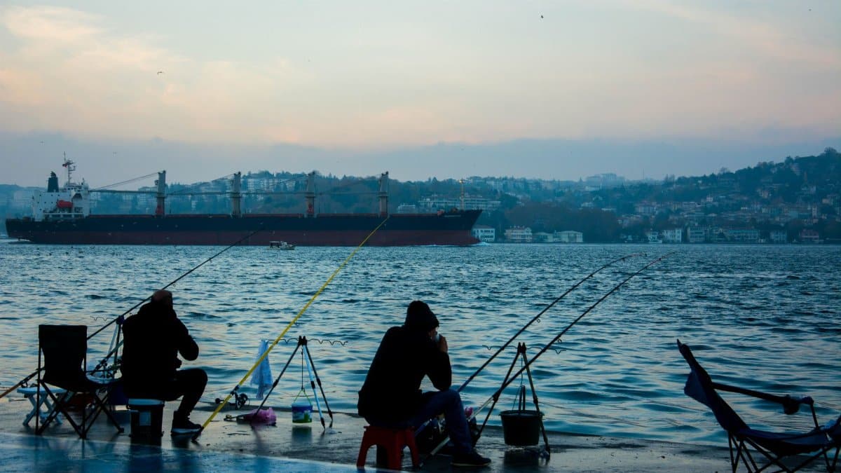 Two men fishing at the harbor during twilight with a large ship in the background. Peaceful and serene scene.